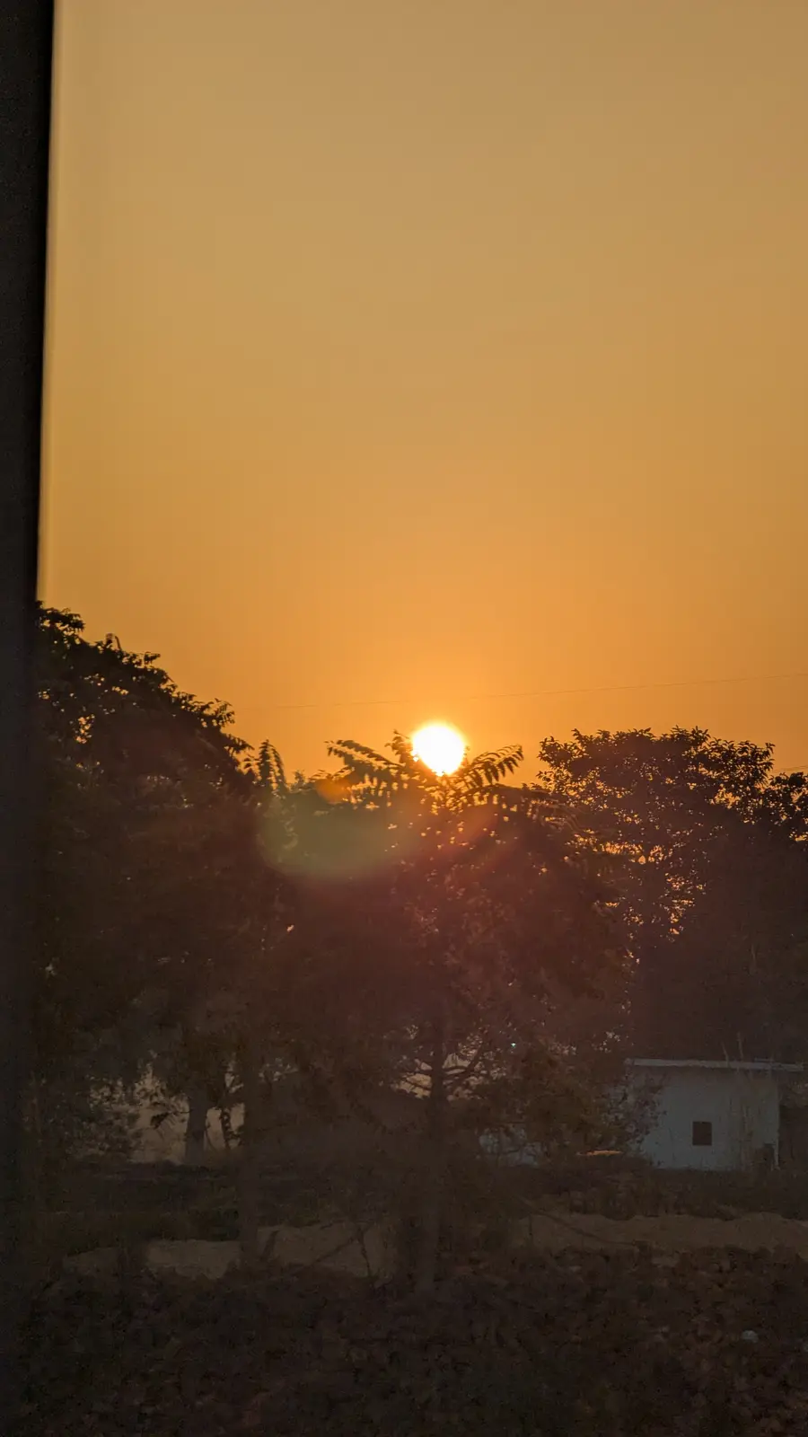 A photograph of a bright orange sunrise viewed through trees, with a small white building visible in the background. The sun…