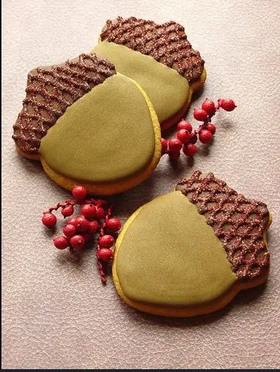 Three acorn-shaped cookies with green icing caps and brown lattice-patterned tops are arranged on a textured surface…
