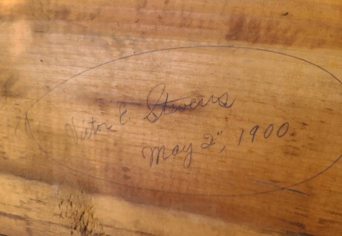 A close-up of a wooden surface with a handwritten signature inside a pencil-drawn oval. The cursive text reads 'Victor E…