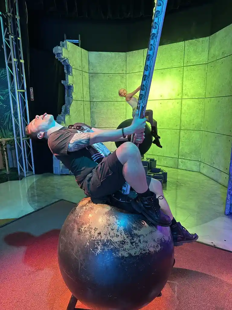 A man with tattoos is lying back on a large, scratched metal ball, gripping a tall pole with a woman clinging to it above…