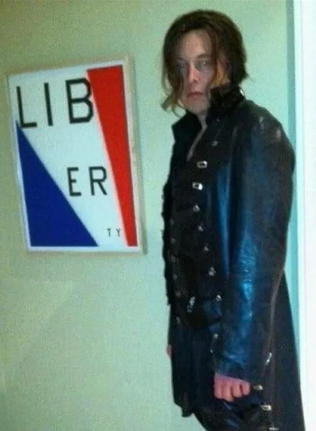 A person in a dark, studded leather outfit stands beside a framed sign with the word 'LIBERTY' split across red and blue…