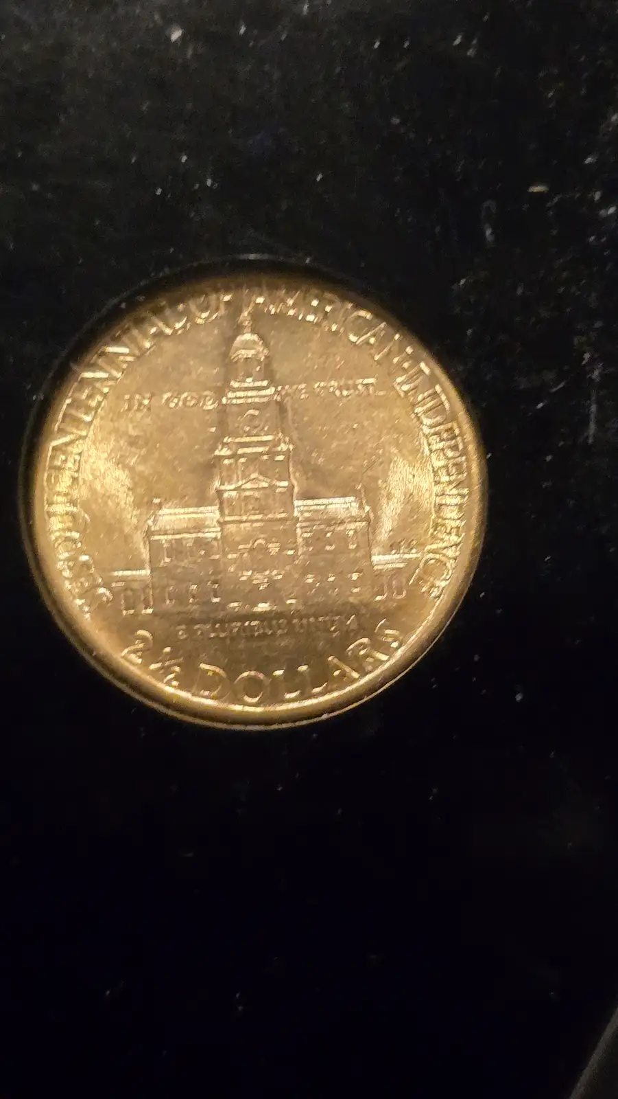 A close-up photo of a gold-colored 2.5-dollar commemorative coin featuring a detailed engraving of a historic building…