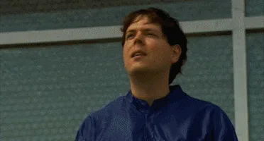 A man in a blue jumpsuit climbs a padded wall, jumps onto a ledge, then onto a giant punching bag suspended in the sky. He…