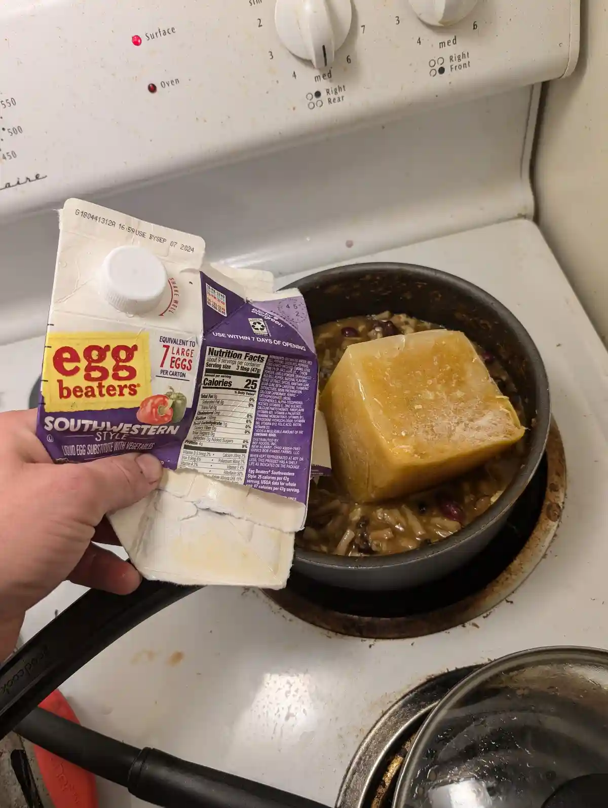 A hand holds a partially opened carton of 'Egg Beaters Southwestern Style' liquid egg substitute over a stovetop, next to a…