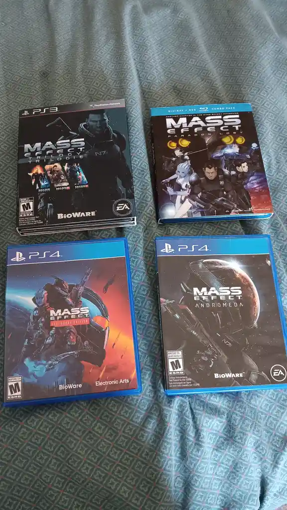 Four Mass Effect video game cases are laid out on a patterned fabric surface: a PS3 'Mass Effect Trilogy' box, a Blu-ray…