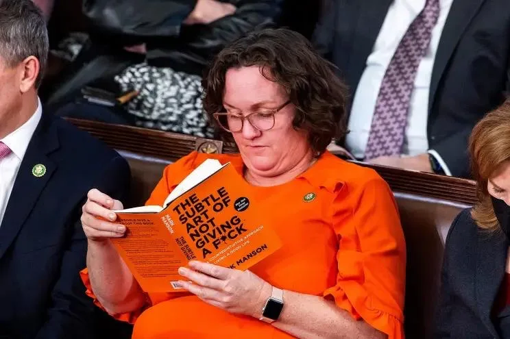 a woman in an orange blouse and glasses is seated in a formal setting, likely a legislative chamber, reading a bright orange…