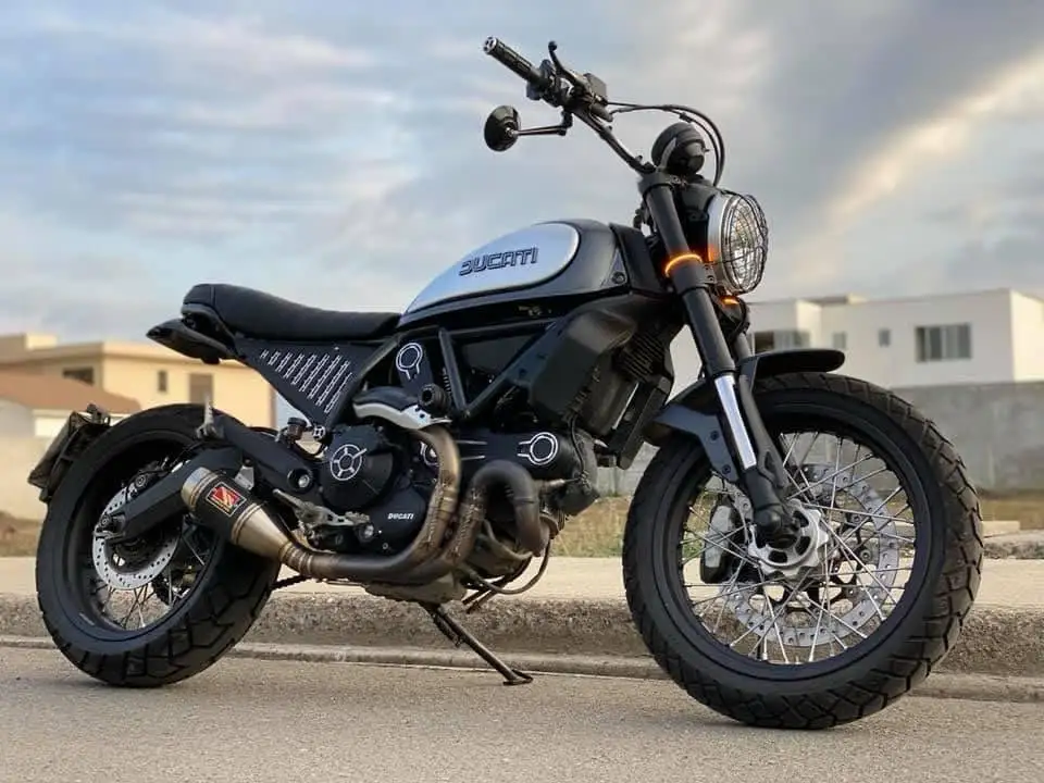 A black Ducati motorcycle with a silver fuel tank and knobby tires is parked on a sidewalk. The bike has a minimalist…