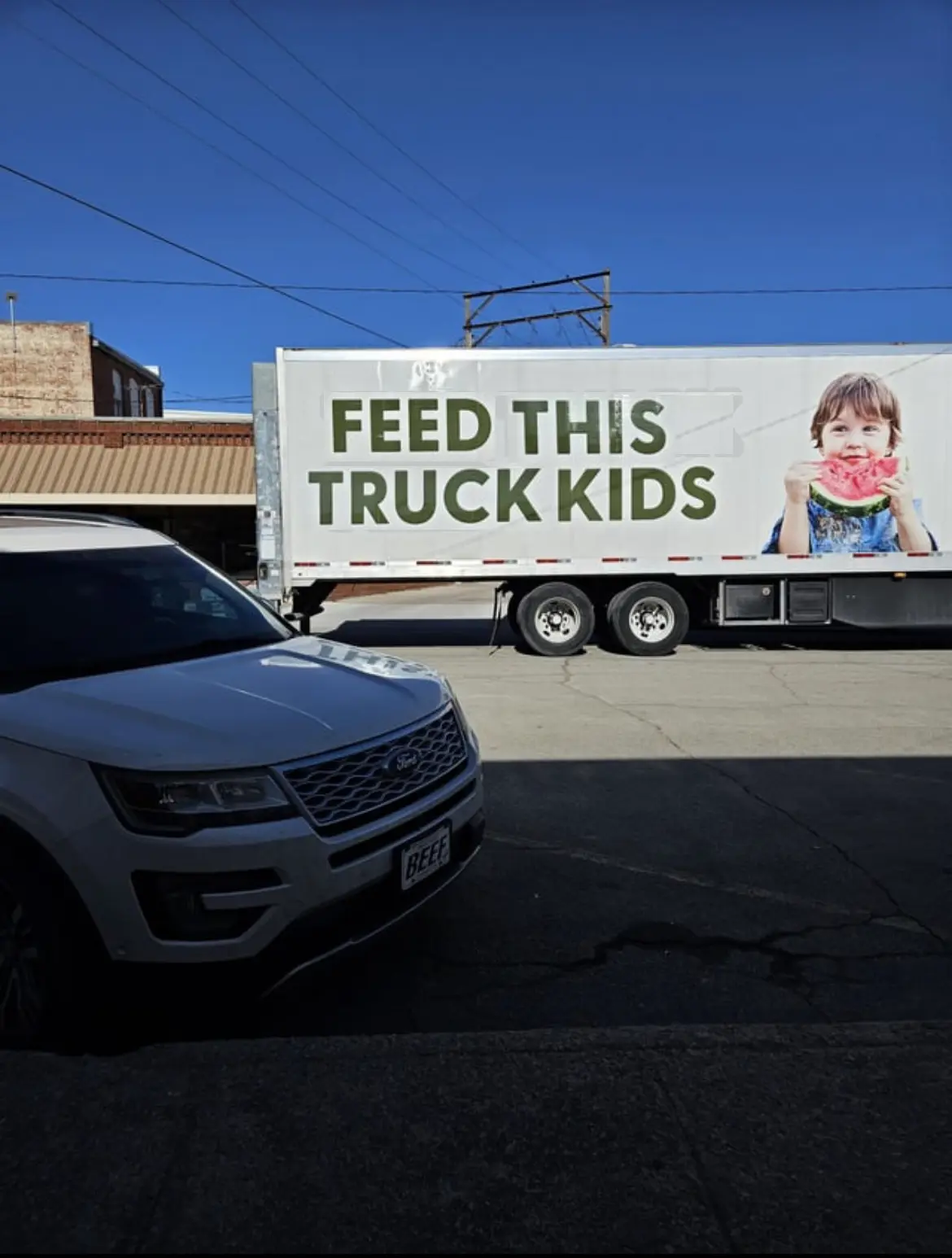 A white semi-truck trailer displays the text 'FEED THIS TRUCK KIDS' alongside an image of a child eating watermelon. In the…