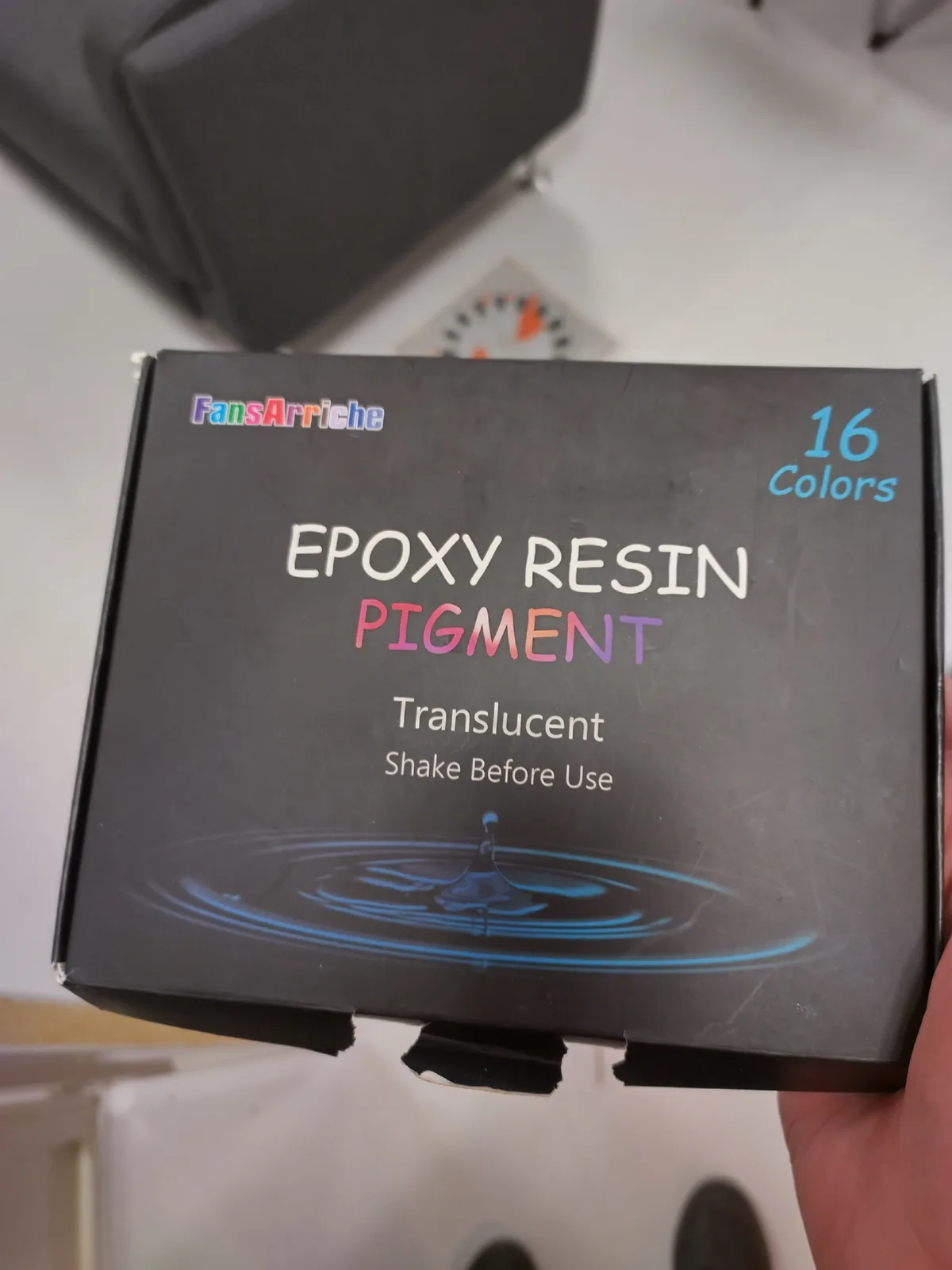 A hand holds a black box labeled 'FansArrieche EPOXY RESIN PIGMENT' with '16 Colors' in the top right corner. The box states…