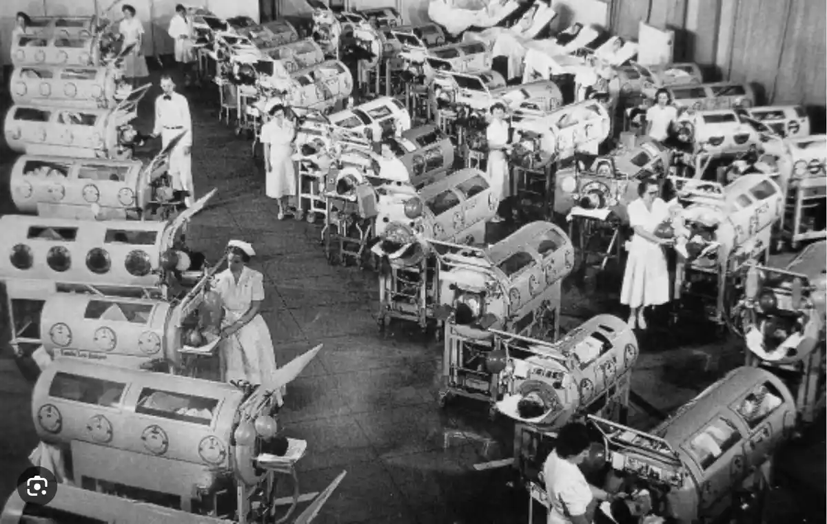 a black-and-white historical photograph shows a large hospital nursery filled with rows of vintage, cylindrical glass and…