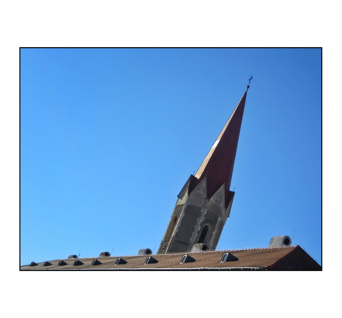 A low-angle photograph shows a church steeple with a red roof and cross against a clear blue sky, rising above a tiled roof…