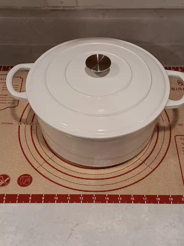 A white ceramic Dutch oven with a lid sits on a beige silicone baking mat marked with red measurement lines and text. The…