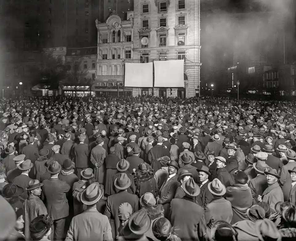 A black-and-white historical photo shows a massive crowd of people, mostly men in hats and coats, gathered at night in front…