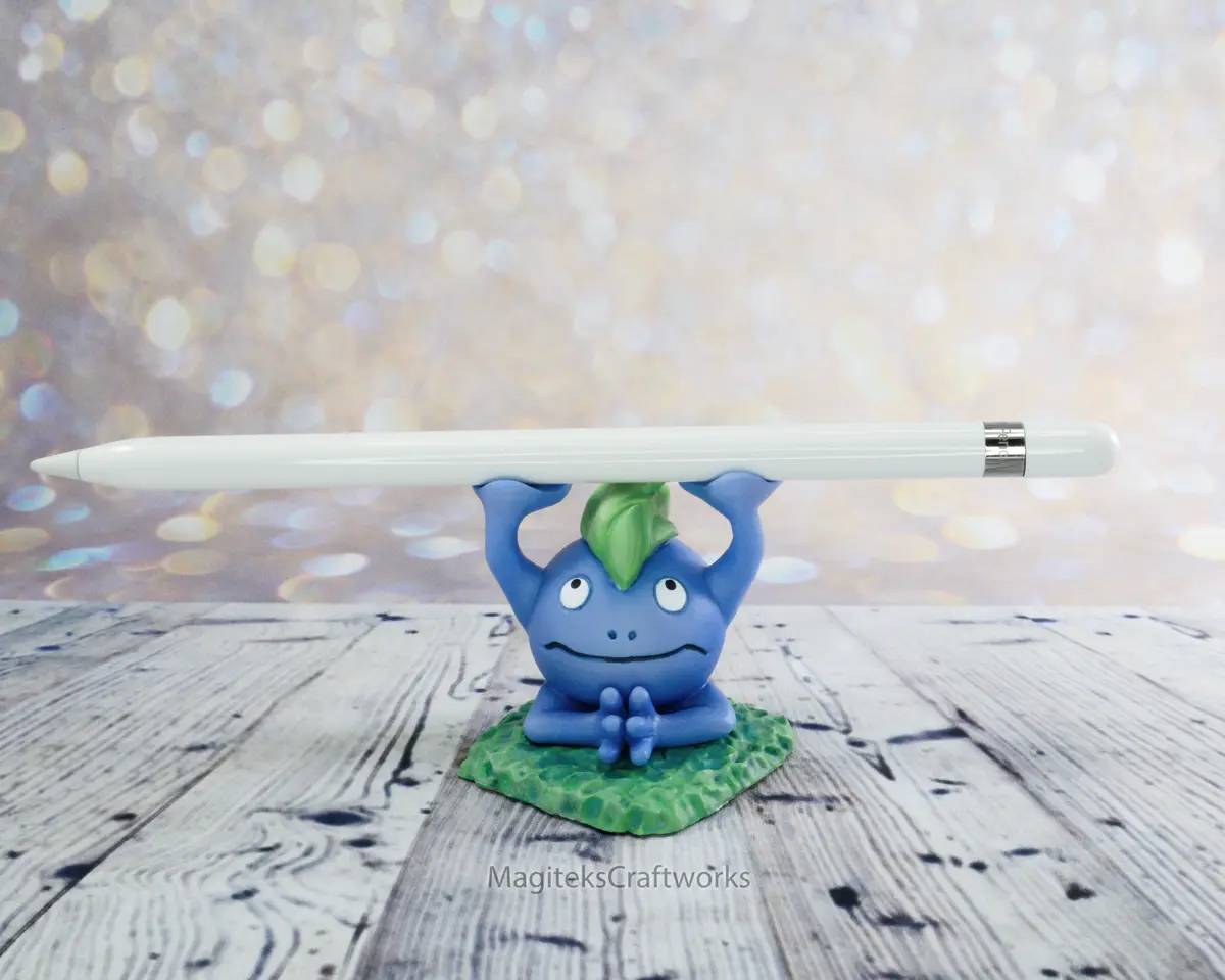 A blue, frog-like Pokémon figurine with a green leaf on its head sits on a leaf-shaped base, holding a white Apple Pencil…