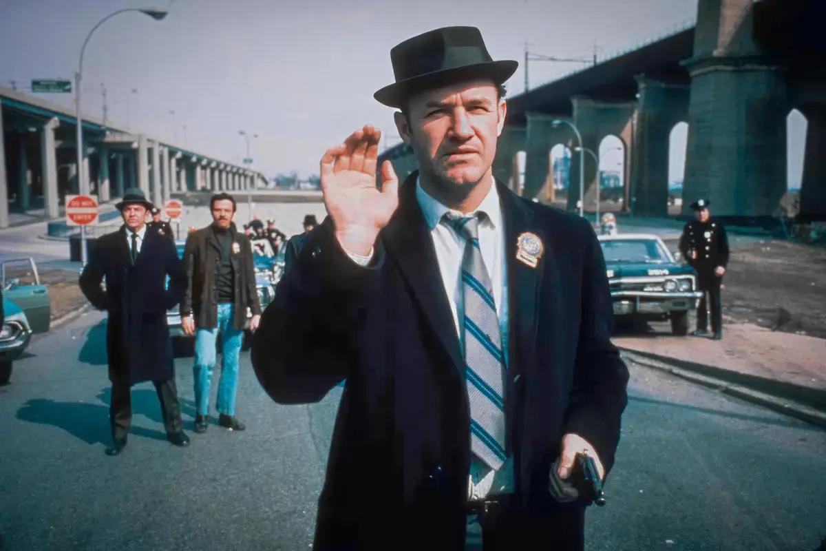 A man in a dark suit, fedora, and tie holds up his hand palm-forward while gripping a handgun in his other hand, standing on…
