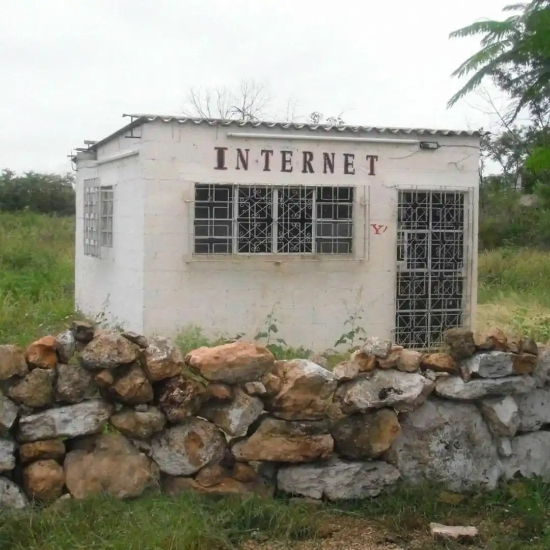A small, white, single-story building with barred windows and a metal roof, labeled 'INTERNET' in large letters, is…