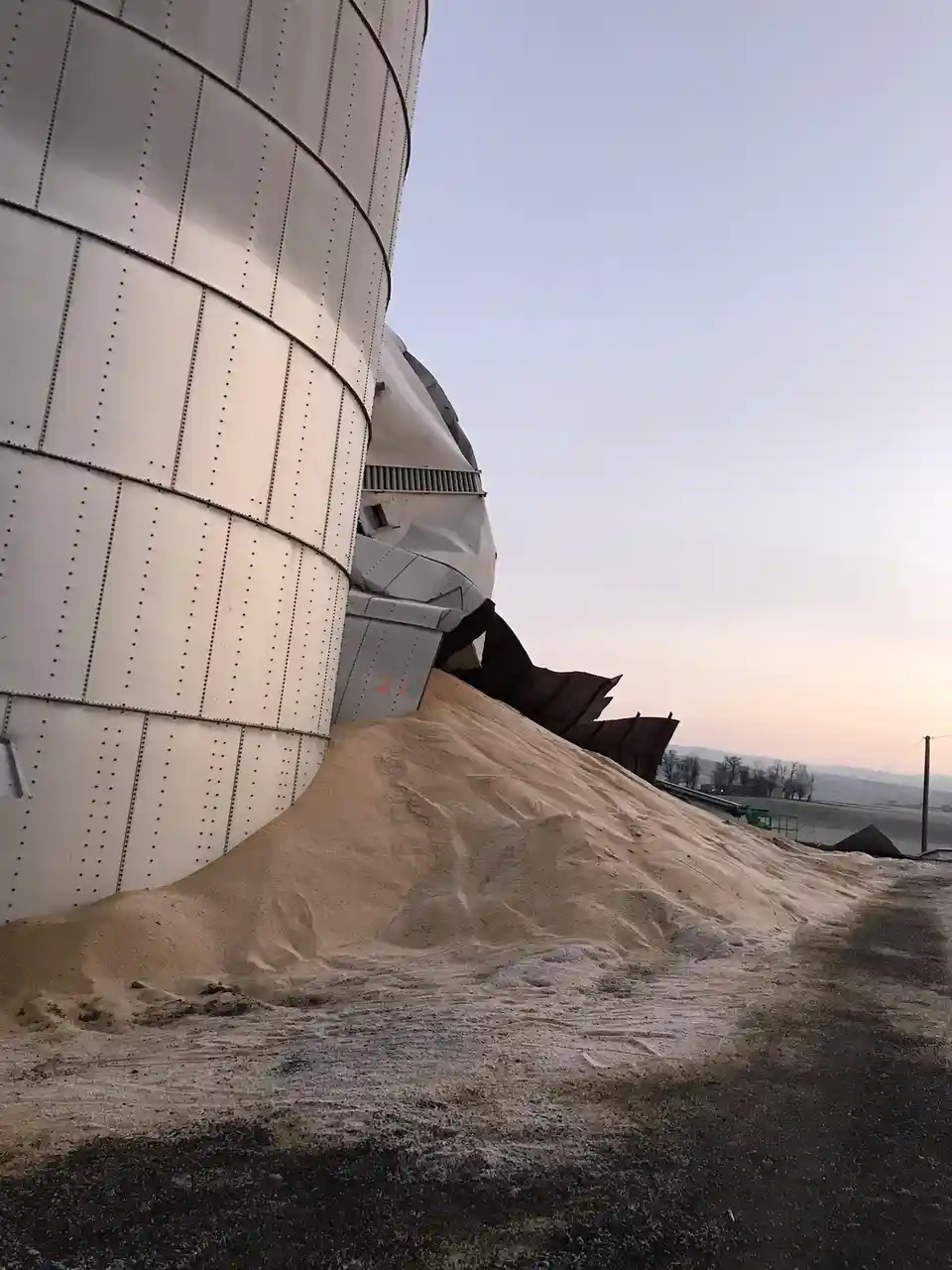 A large, white, riveted metal silo is visibly damaged, with its side panels torn open and a massive pile of sand spilling…