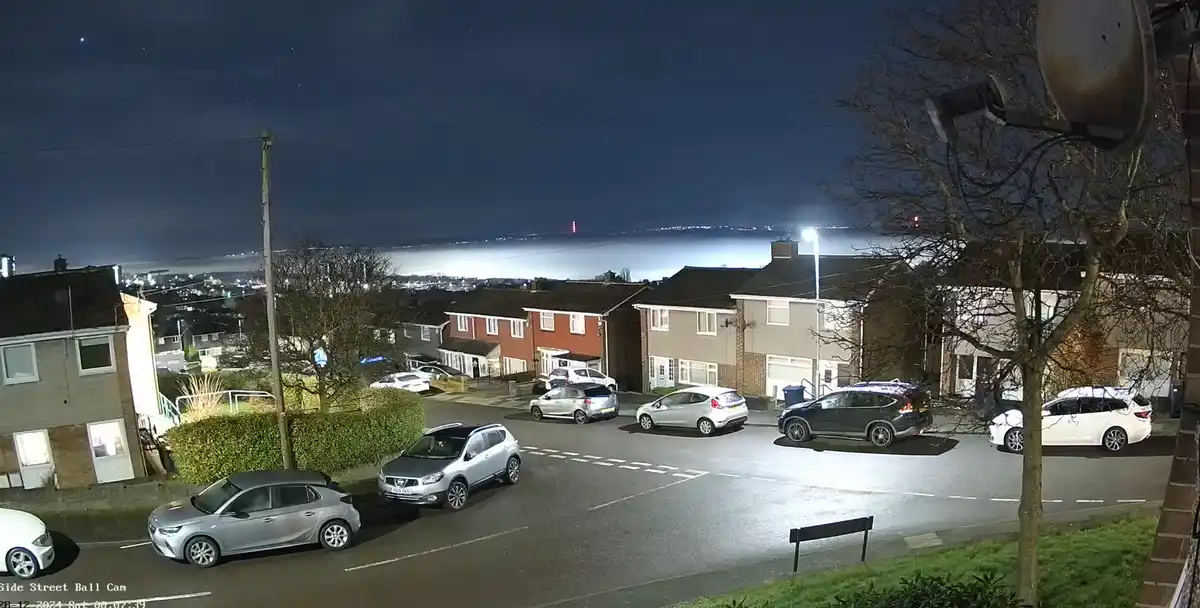 A nighttime security camera view of a residential street with parked cars, houses, and a distant cityscape under a starry…