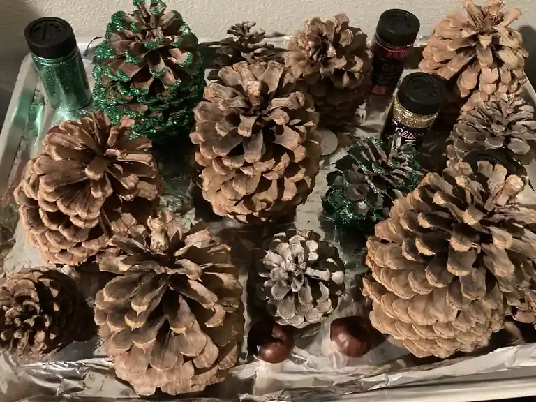 A tray lined with aluminum foil holds numerous pine cones of various sizes, some decorated with green glitter, alongside…