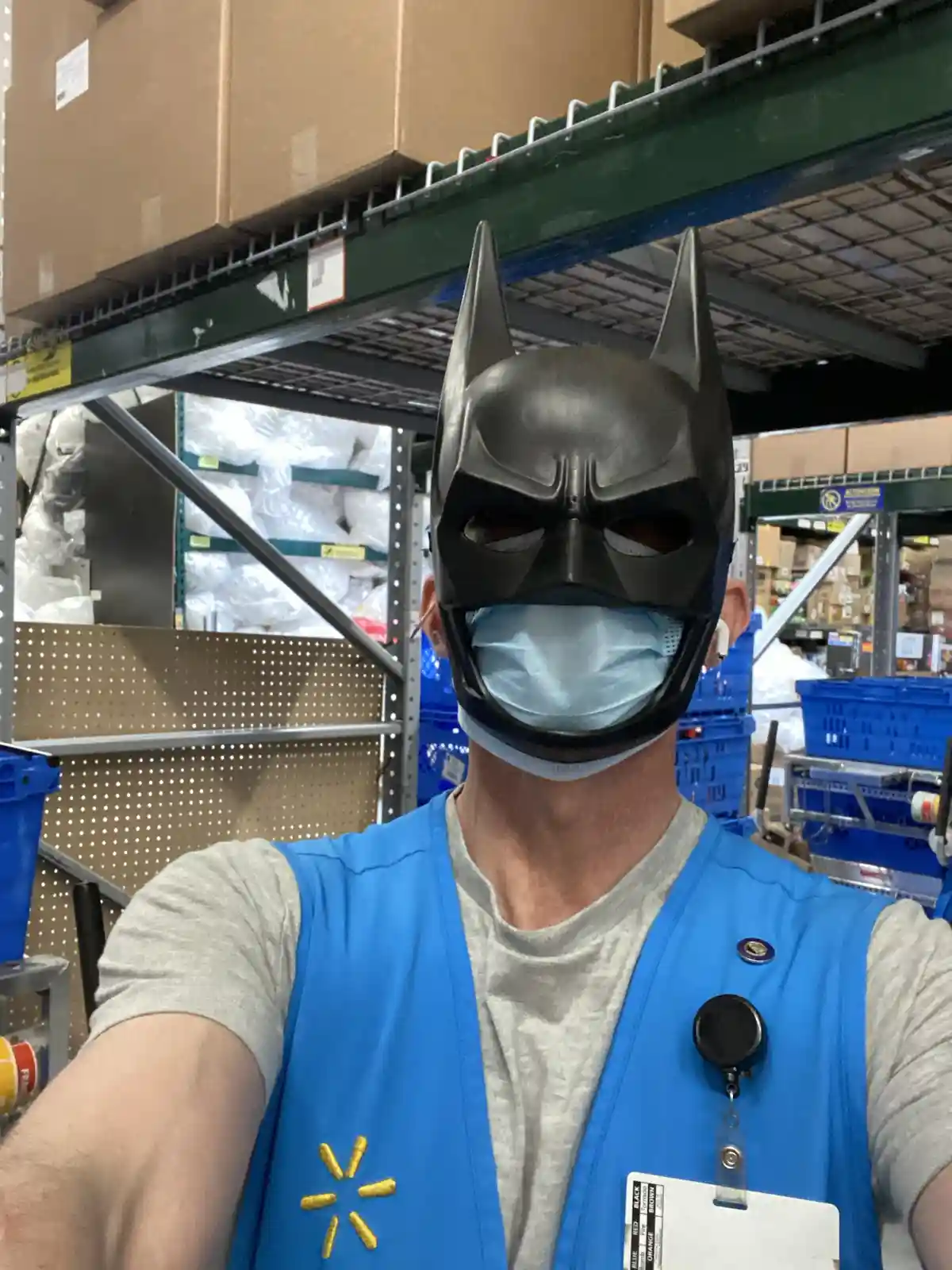 A person wearing a blue Walmart vest and a light gray t-shirt takes a selfie in a warehouse aisle, with tall shelves of…