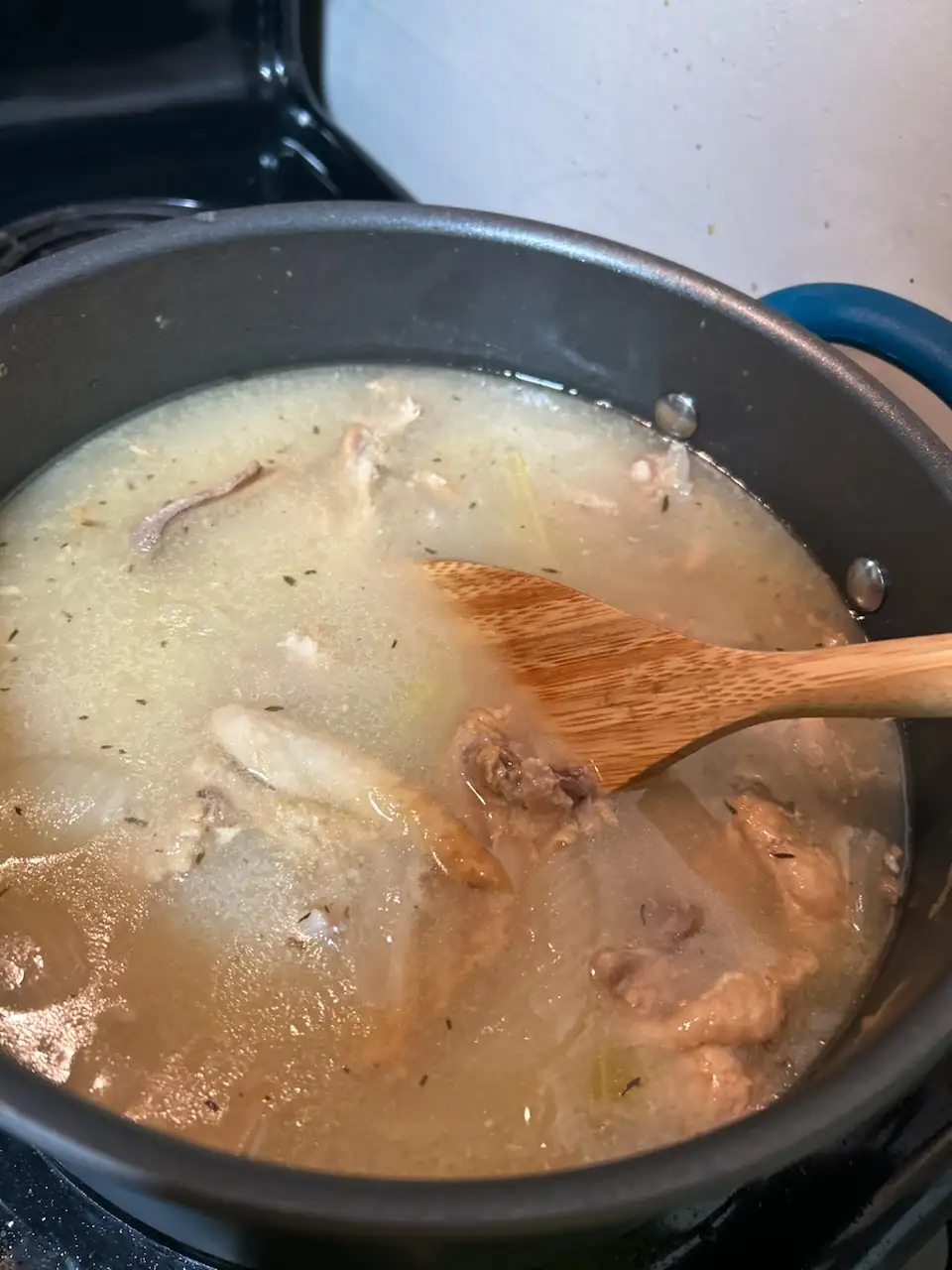 A close-up photo shows a dark pot on a stovetop filled with a cloudy, light-brown broth containing visible meat bones…