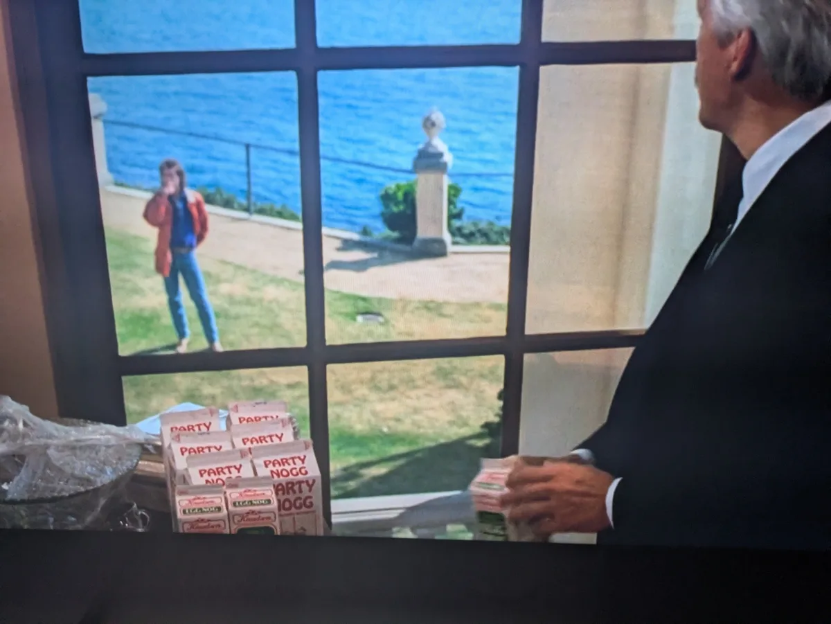 A man in a suit sits indoors at a desk with multiple boxes of 'Party Nogg' drink cartons, looking out a window at a young…