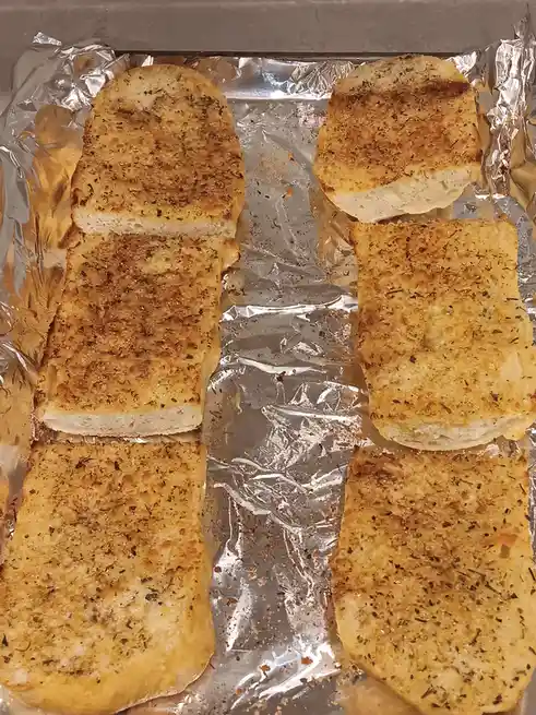 Six slices of garlic bread, golden-brown and seasoned with herbs, are arranged on a baking sheet lined with aluminum foil…