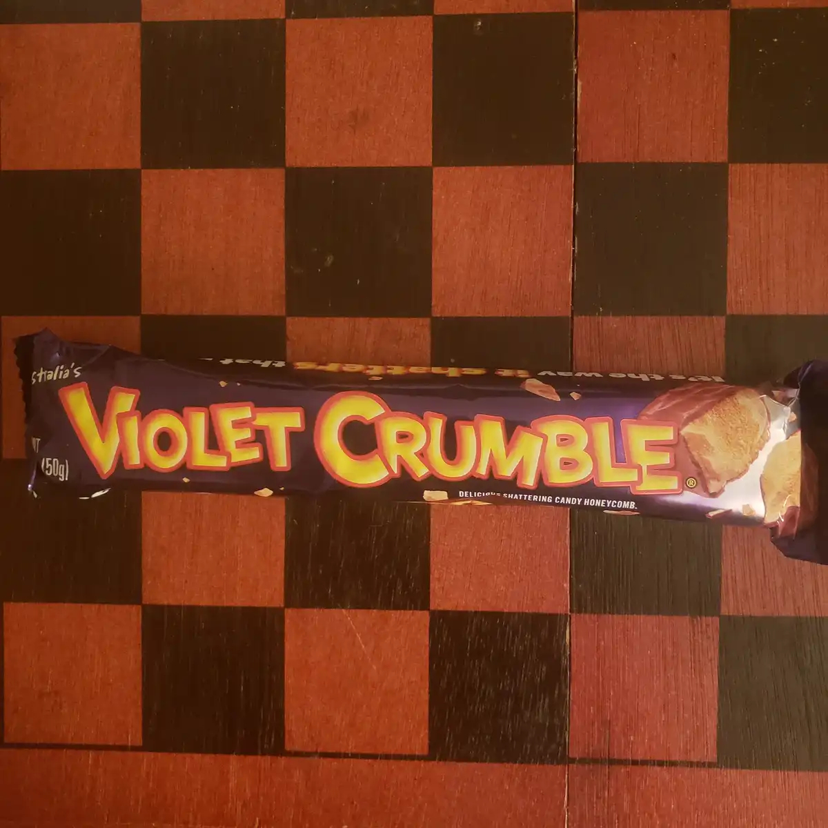 A purple-wrapped Violet Crumble chocolate bar lies horizontally on a red and black checkered wooden surface. The packaging…