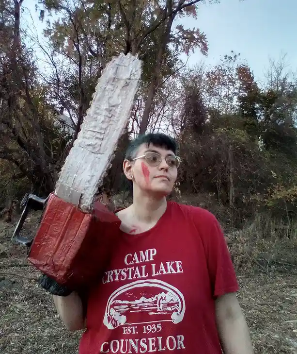 A person with short dark hair and glasses wears a red t-shirt that reads 'CAMP CRYSTAL LAKE EST. 1935 COUNSELOR' and carries…