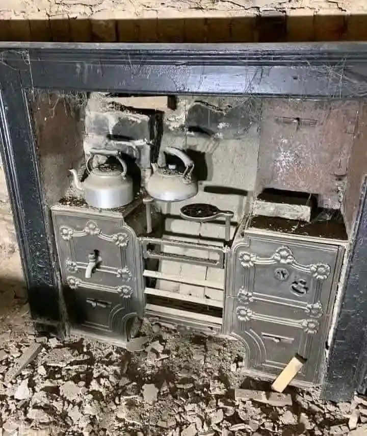 A dilapidated, ornate cast-iron fireplace insert with two silver kettles sits amidst rubble and debris, with the overlaid…