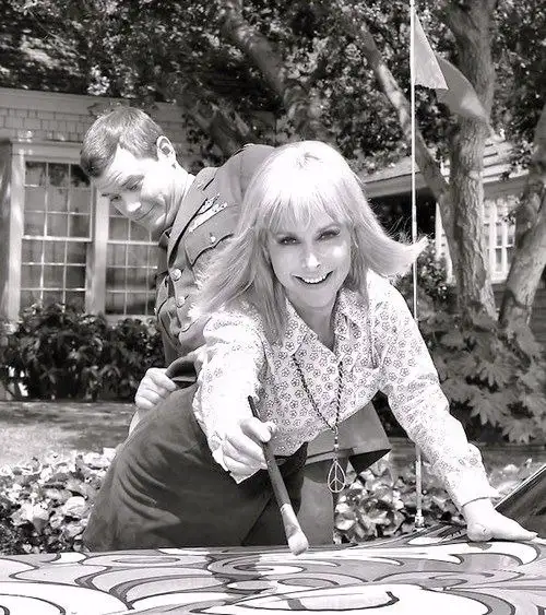 A black-and-white photo shows a smiling blonde woman in a patterned blouse leaning over a car with a psychedelic paint job…