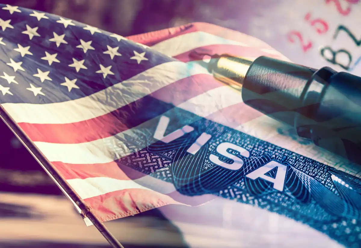 A close-up image shows an American flag waving in the foreground, with a U.S. visa document partially visible underneath…