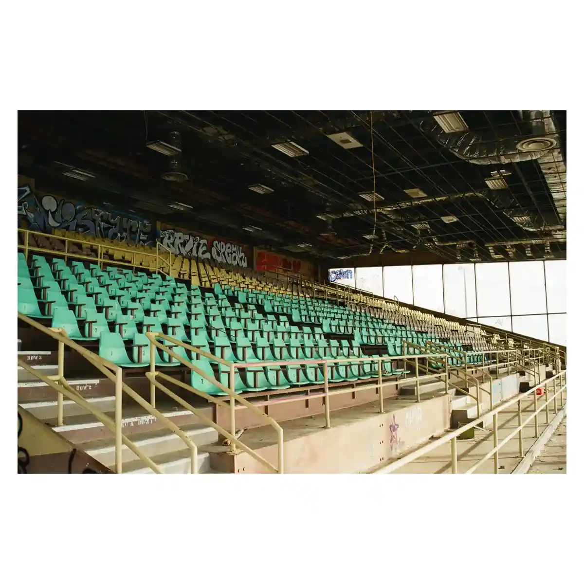 An empty, dilapidated stadium with rows of faded green and yellow seats, graffiti-covered walls and ceiling, and visible…