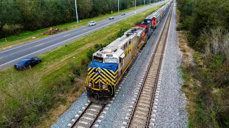 An aerial photo shows a blue and yellow train on tracks running parallel to a road. A dark blue car is parked on the grassy…