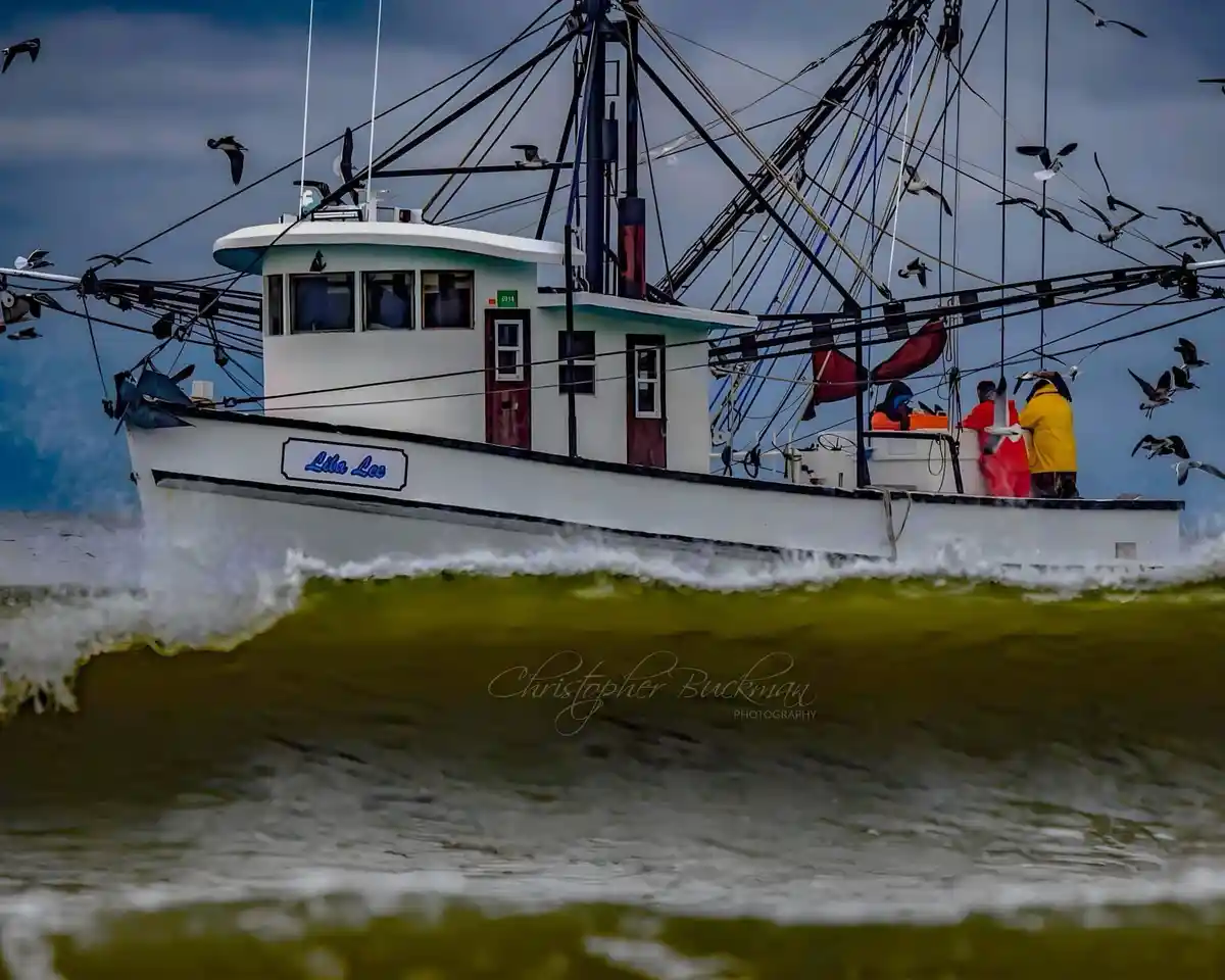 A fishing boat named 'Lila Lee' cuts through choppy, greenish water, with several crew members in bright jackets visible on…