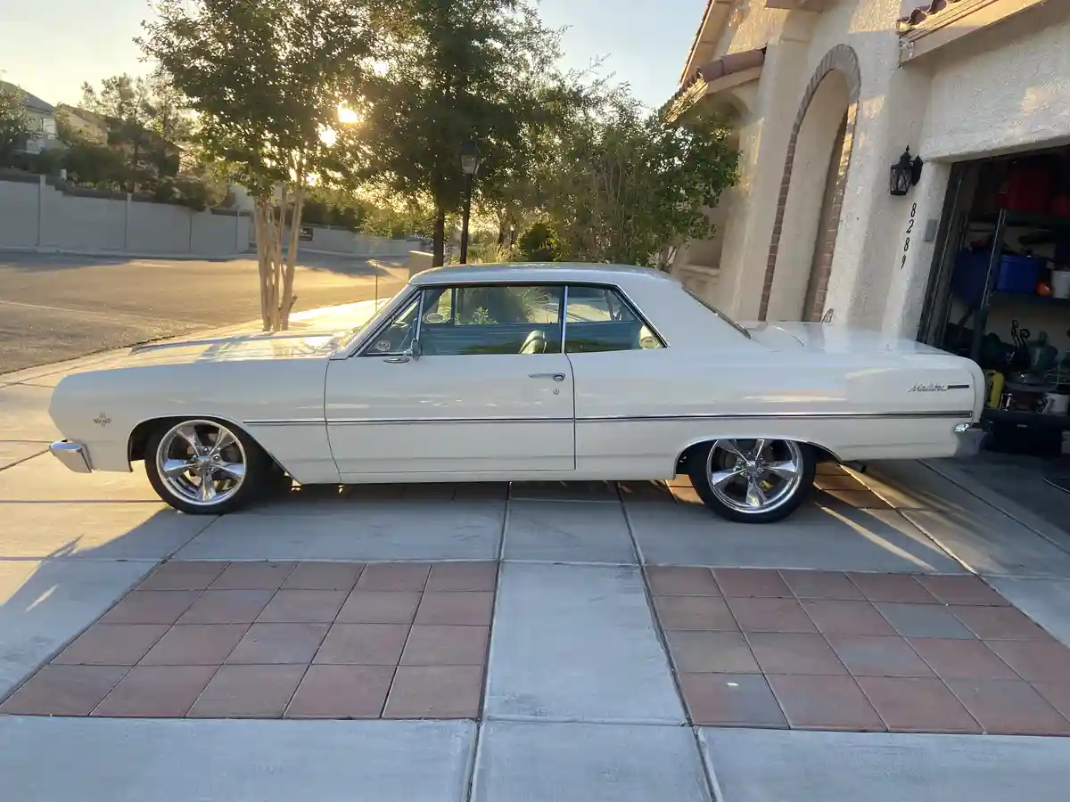 A white 1960s Chevrolet Malibu classic car is parked on a tiled driveway in front of a house, with the sun setting behind…