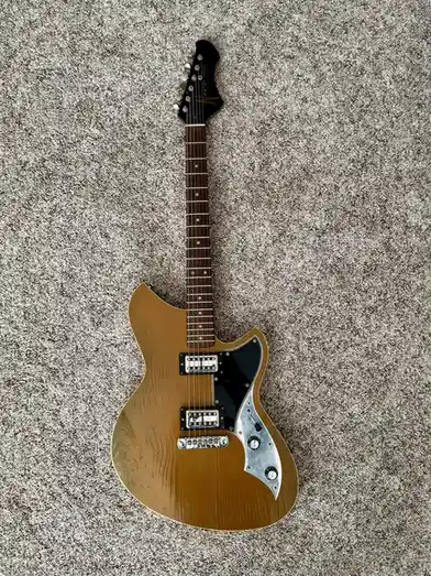 A light brown electric guitar with a black pickguard and chrome hardware lies on a gray carpet. The overlaid text reads 'i…