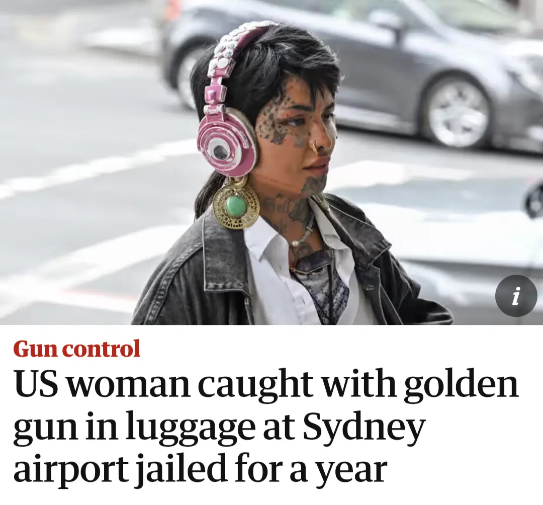 A person with tattoos, a nose ring, and large pink headphones stands on a street with a car blurred in the background…
