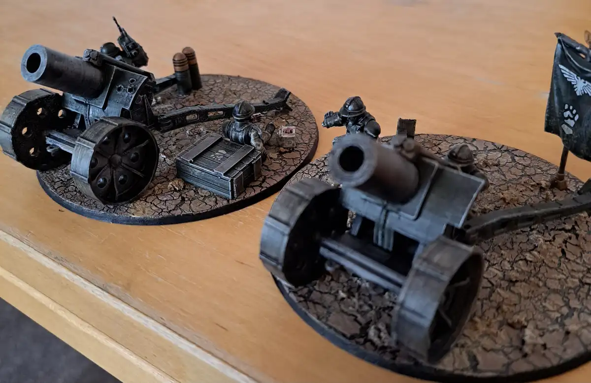Two miniature artillery pieces on cracked-brown circular bases sit on a wooden table, one in the foreground and one behind…