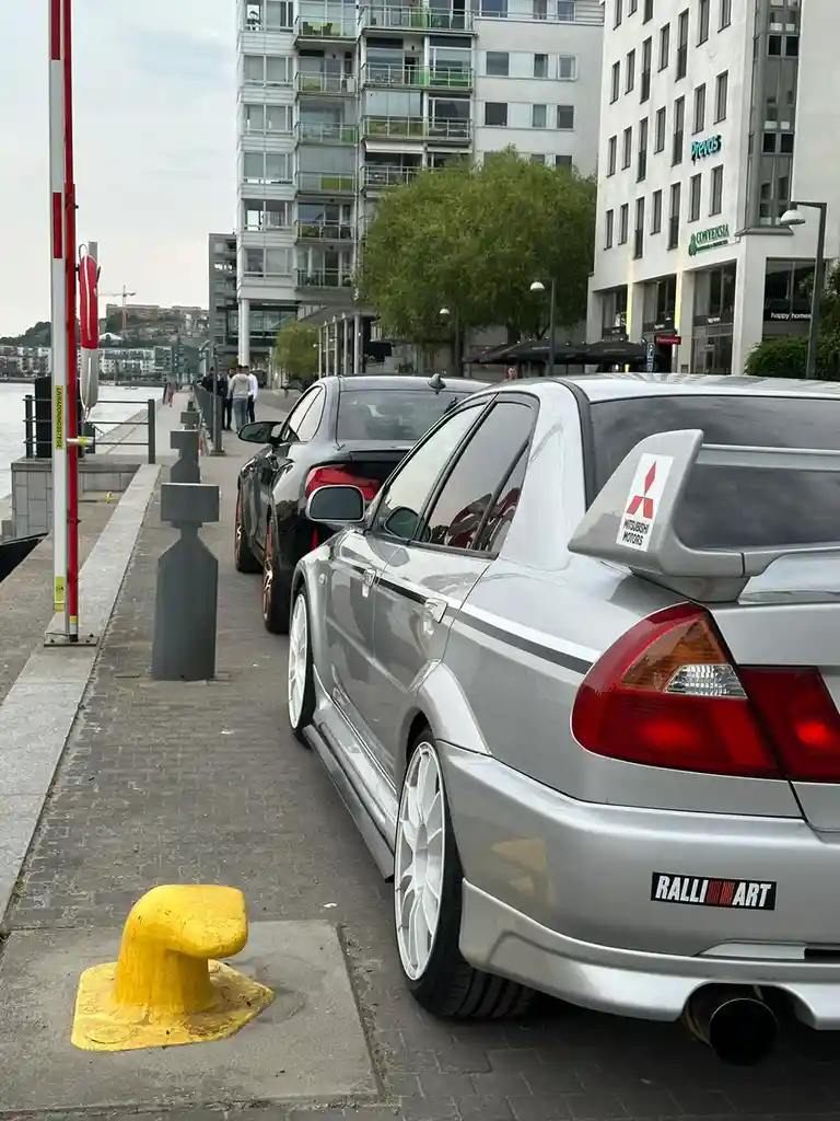 A silver Mitsubishi Lancer Evolution with a Ralliart badge parked on a city sidewalk next to a yellow bollard, with a black…