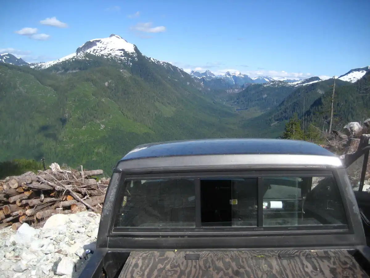 A black pickup truck with a wooden bed liner is parked on a rocky overlook, facing a vast, scenic mountain range with…