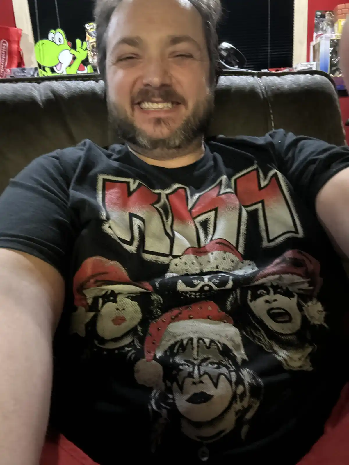 A man with a beard and mustache is smiling broadly while sitting on a couch, wearing a black t-shirt with the band KISS and…