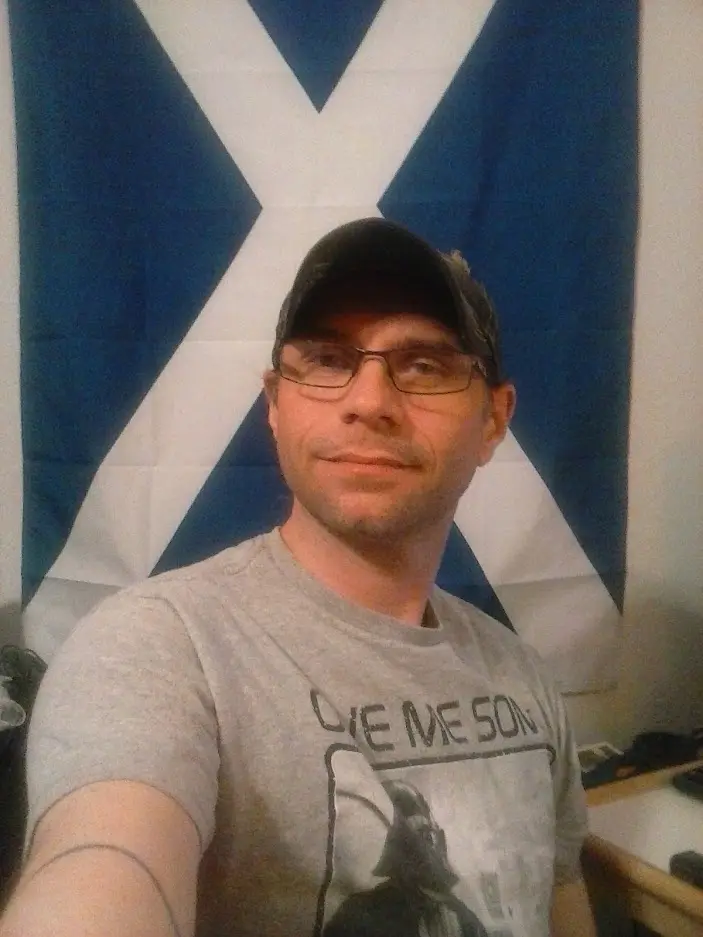 A man wearing glasses and a baseball cap takes a selfie in front of a Scottish flag. He wears a gray t-shirt with the text…