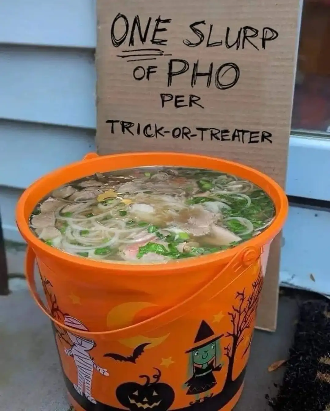 An orange Halloween-themed bucket filled with pho soup sits outdoors, with a cardboard sign behind it that reads 'ONE SLURP…