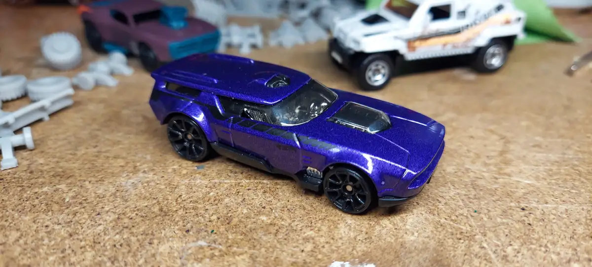 A shiny purple toy car with black wheels sits on a wooden surface, surrounded by other toy cars and white plastic parts. The…