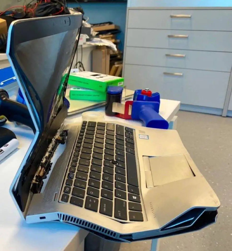 A laptop is shown with its screen severely bent backward, exposing internal components, sitting on a cluttered desk with…