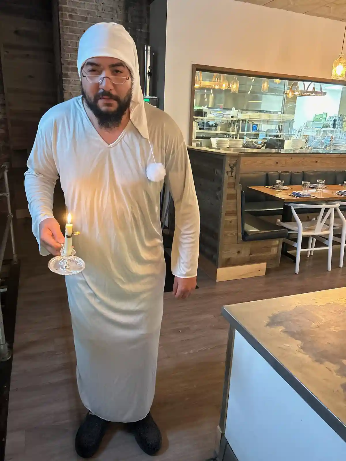 A man with a beard and glasses stands in a restaurant wearing a white robe and head covering resembling a Santa hat with a…