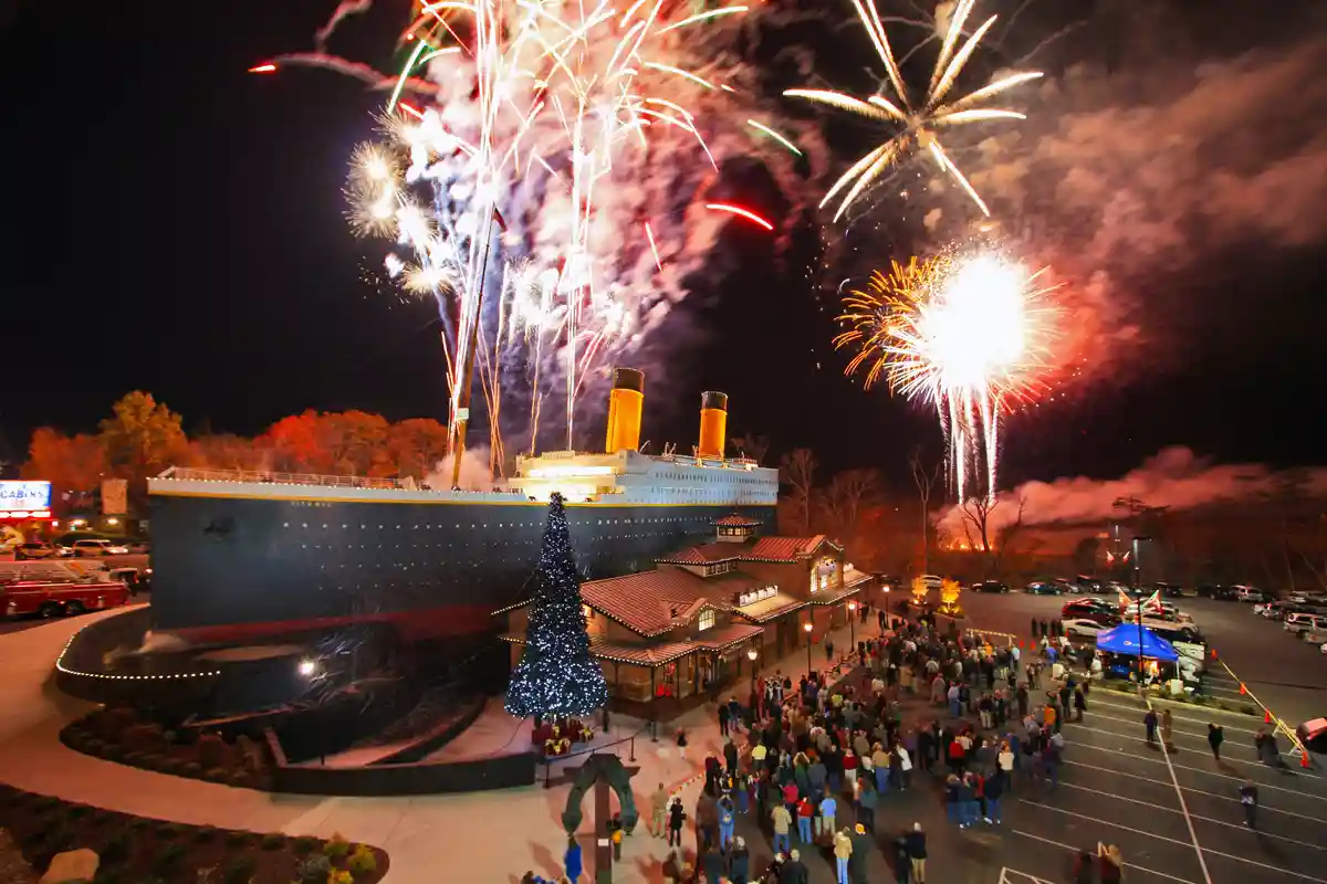 A nighttime photo shows a large, stationary replica of the Titanic ship with fireworks exploding above it, while crowds of…