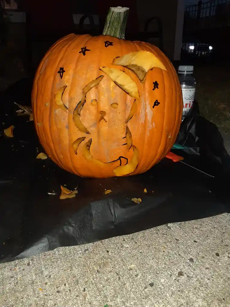 A large orange pumpkin sits on a black plastic sheet outdoors at night, its surface carved with a crude, asymmetrical face…
