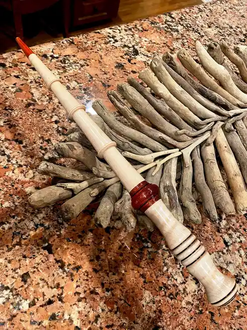 A polished wooden flute with red accents lies diagonally across a pile of weathered driftwood arranged to resemble a…
